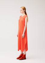 A woman with long, wavy hair in a loose ponytail stands sideways against a plain light background, wearing the MONTHLY COLORS : SEPTEMBER sleeveless, ankle-length orange dress and matching orange high-top shoes by PLEATS PLEASE ISSEY MIYAKE.
