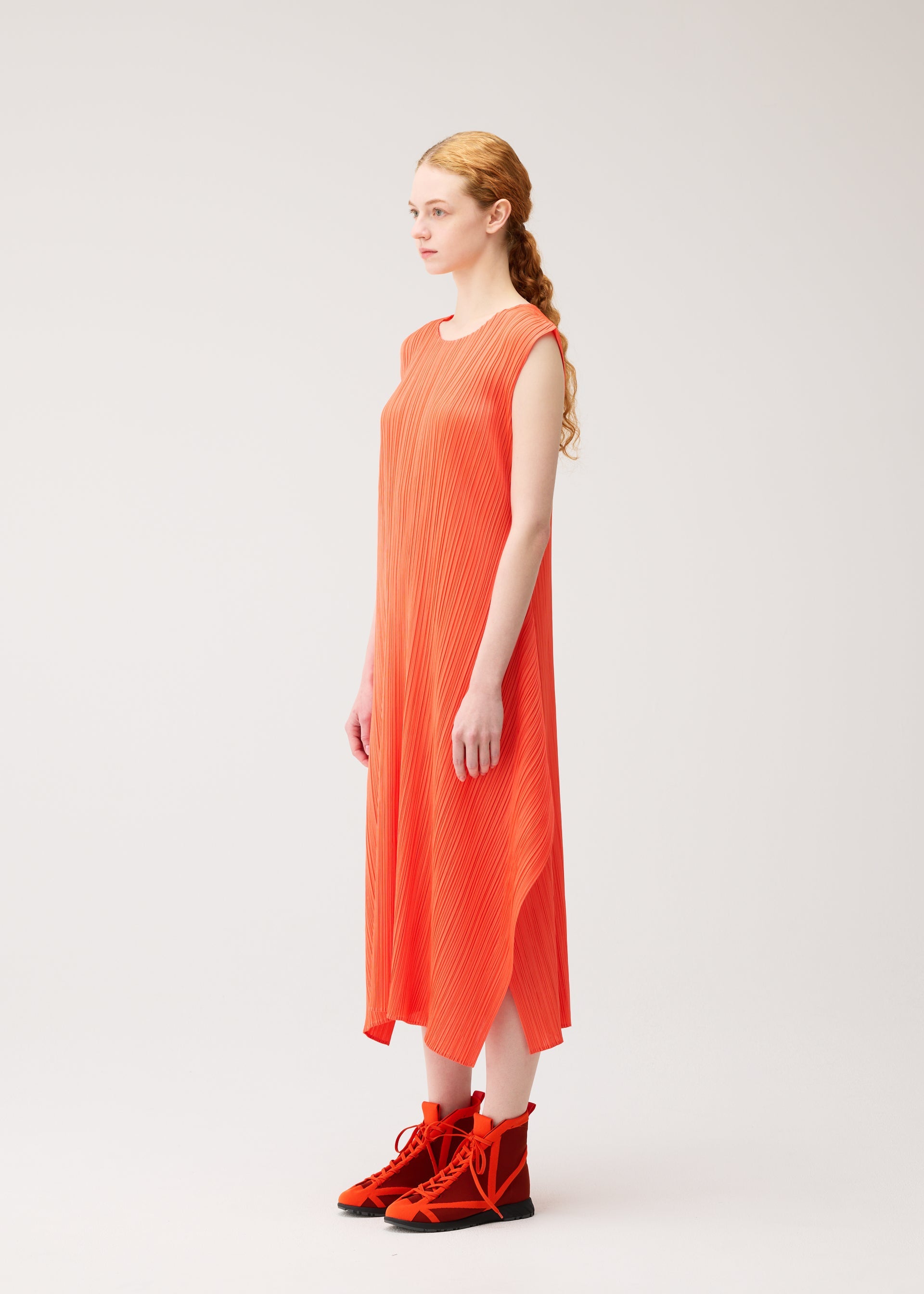 Zoomed view A woman with long, wavy hair in a loose ponytail stands sideways against a plain light background, wearing the MONTHLY COLORS : SEPTEMBER sleeveless, ankle-length orange dress and matching orange high-top shoes by PLEATS PLEASE ISSEY MIYAKE.