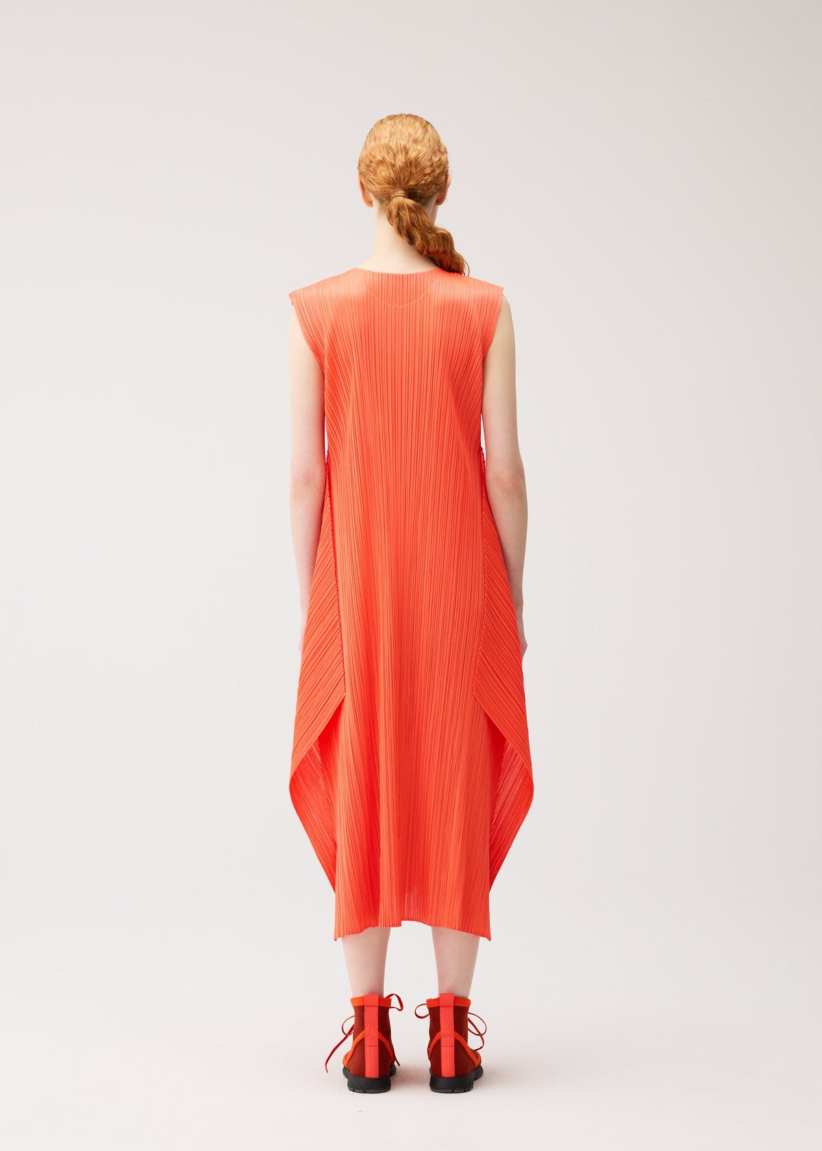 A person with long, braided hair wears the MONTHLY COLORS : SEPTEMBER sleeveless bright orange pleated midi dress by PLEATS PLEASE ISSEY MIYAKE—styled with a round neck and flared silhouette—and matching orange sandals, standing against a plain light background, facing away from the camera. view 9 of 11