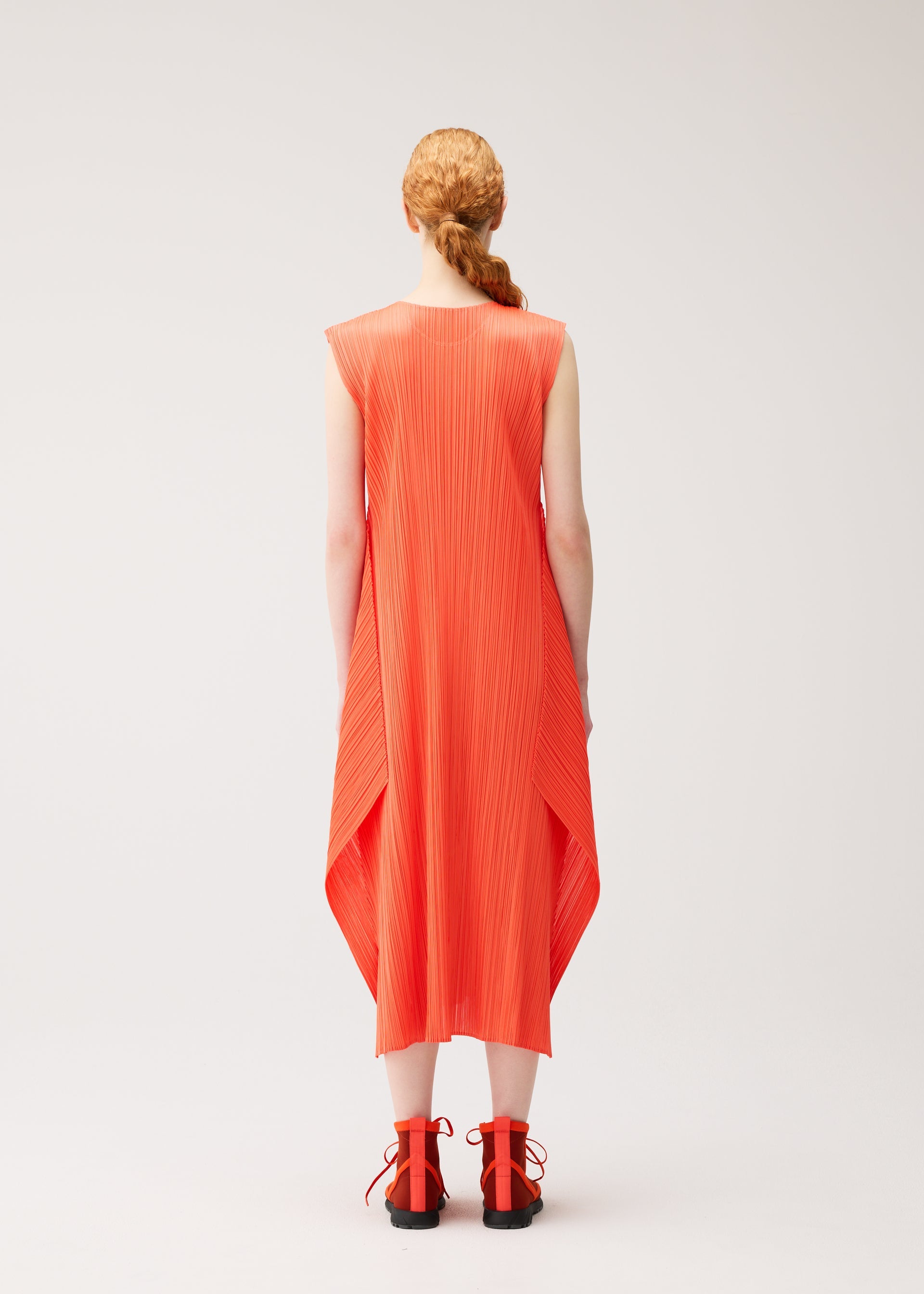 Zoomed view A person with long, braided hair wears the MONTHLY COLORS : SEPTEMBER sleeveless bright orange pleated midi dress by PLEATS PLEASE ISSEY MIYAKE—styled with a round neck and flared silhouette—and matching orange sandals, standing against a plain light background, facing away from the camera.