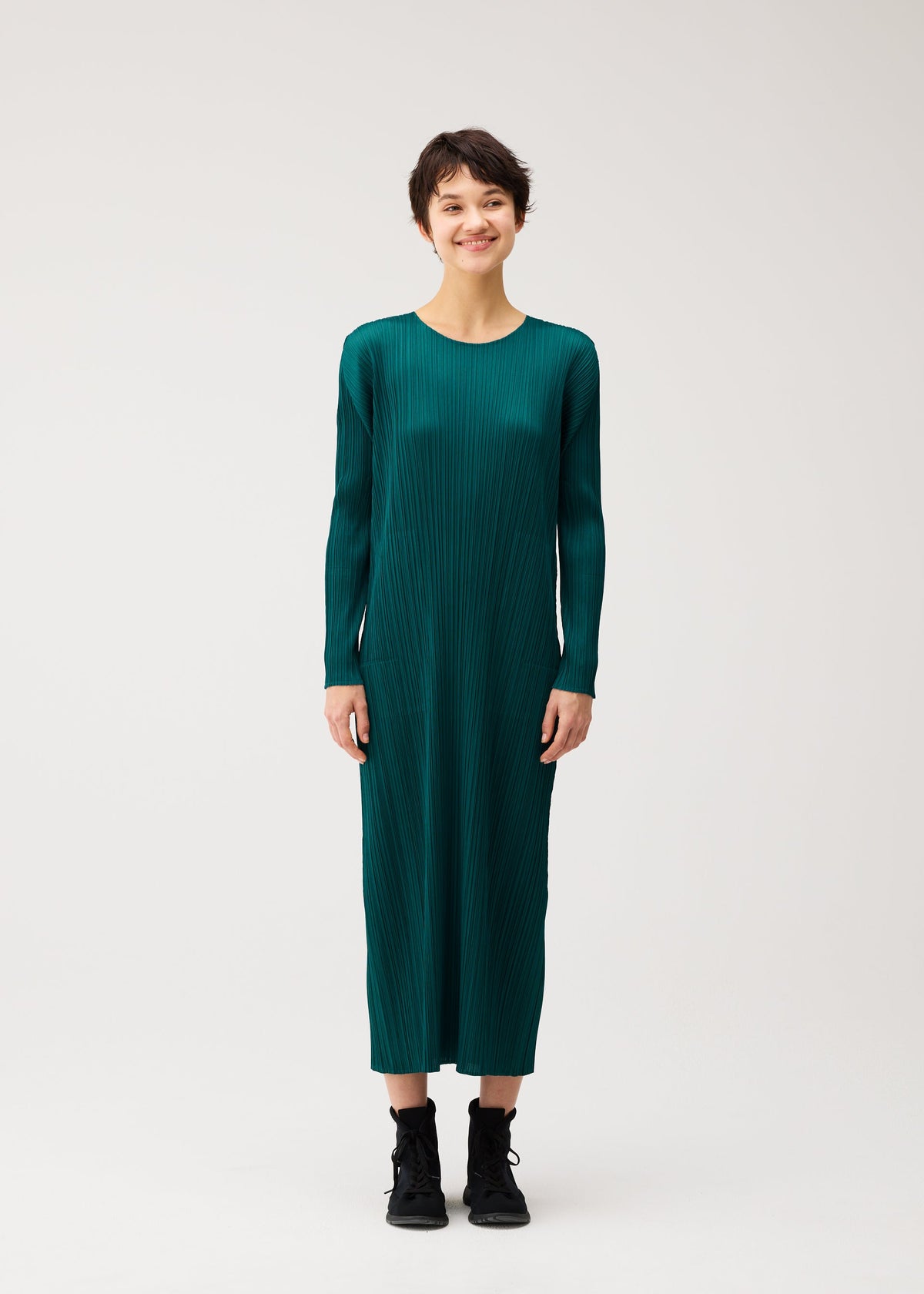 A person with short dark hair smiles in the MONTHLY COLORS : OCTOBER dress by PLEATS PLEASE ISSEY MIYAKE—green, long-sleeved, pleated with pockets—paired with black boots against a plain light background. view 6 of 9