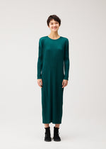 A person with short dark hair smiles in the MONTHLY COLORS : OCTOBER dress by PLEATS PLEASE ISSEY MIYAKE—green, long-sleeved, pleated with pockets—paired with black boots against a plain light background.