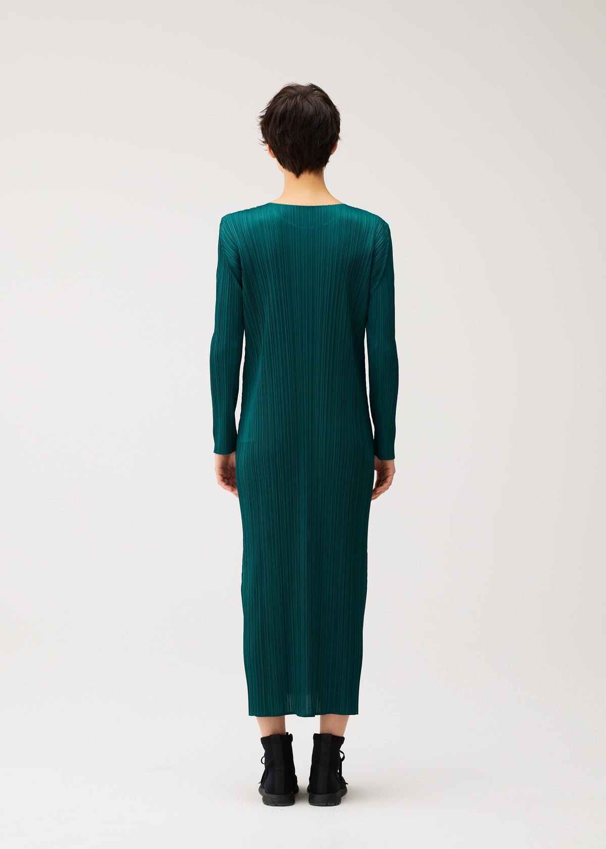 A person with short dark hair faces away, wearing the MONTHLY COLORS : OCTOBER long-sleeved, teal pleated dress by PLEATS PLEASE ISSEY MIYAKE and black ankle boots, against a plain light background. view 8 of 9