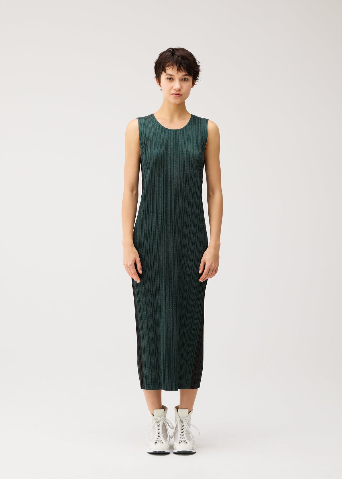 Zoomed view A person with short dark hair wears the COTTON DENIM by PLEATS PLEASE ISSEY MIYAKE, a sleeveless dark green ribbed dress, paired with white lace-up boots against a plain light background.