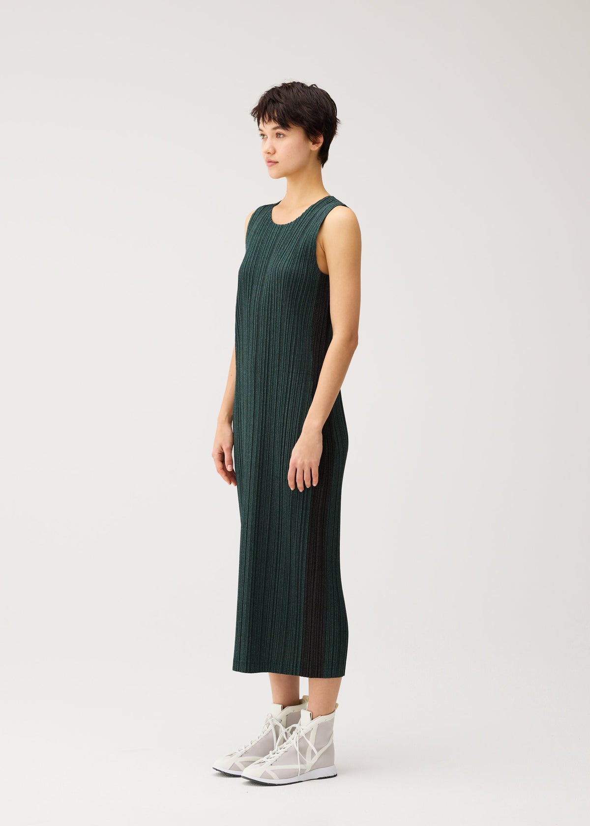 Zoomed view A person with short dark hair wears the PLEATS PLEASE ISSEY MIYAKE COTTON DENIM sleeveless, calf-length dress, paired with white high-top sneakers, standing sideways against a plain white background.