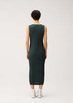 A person with short dark hair wears the PLEATS PLEASE ISSEY MIYAKE COTTON DENIM sleeveless dress in ribbed dark green, paired with white sneakers, standing against a plain light background.