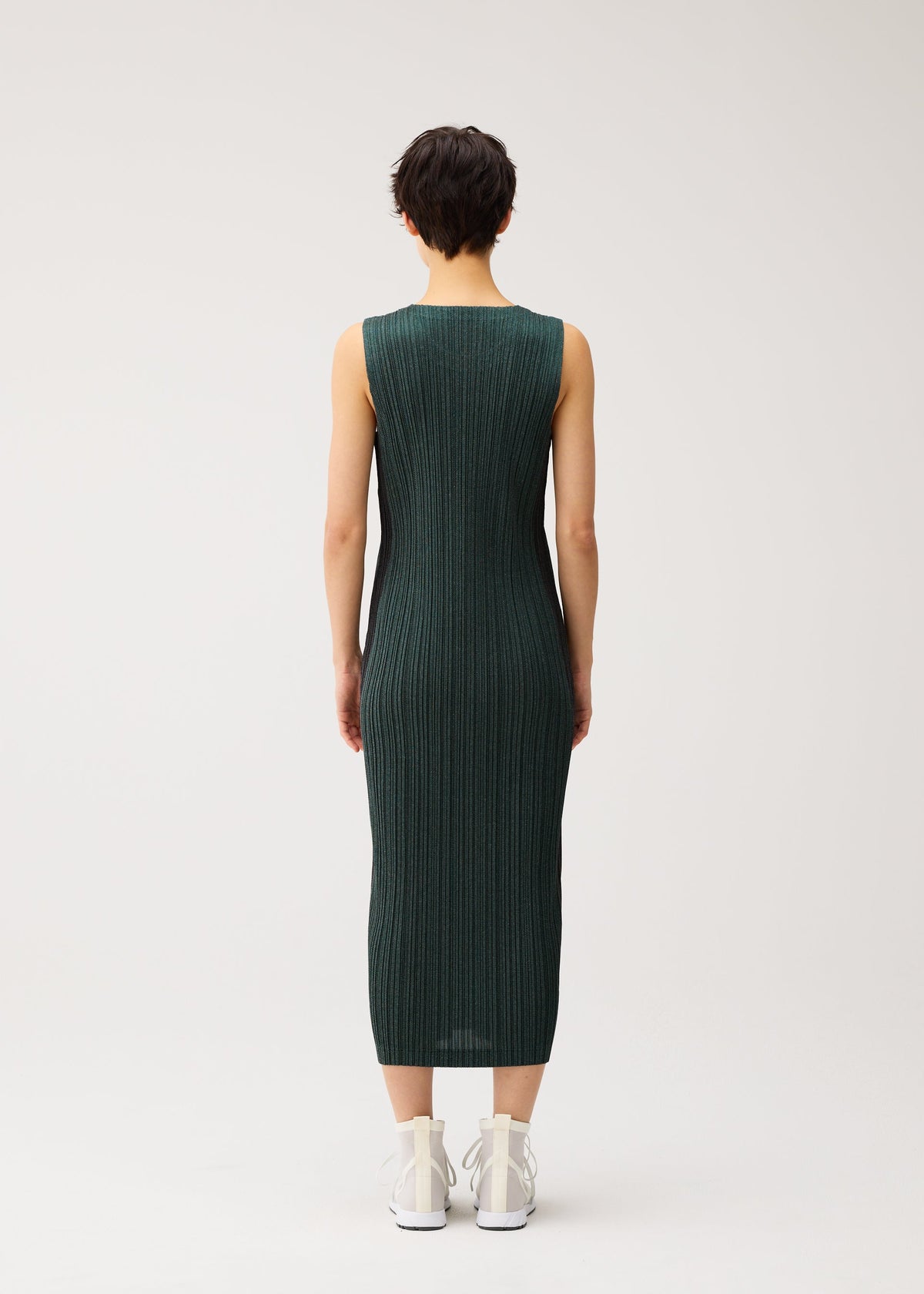 Zoomed view A person with short dark hair wears the PLEATS PLEASE ISSEY MIYAKE COTTON DENIM sleeveless dress in ribbed dark green, paired with white sneakers, standing against a plain light background.