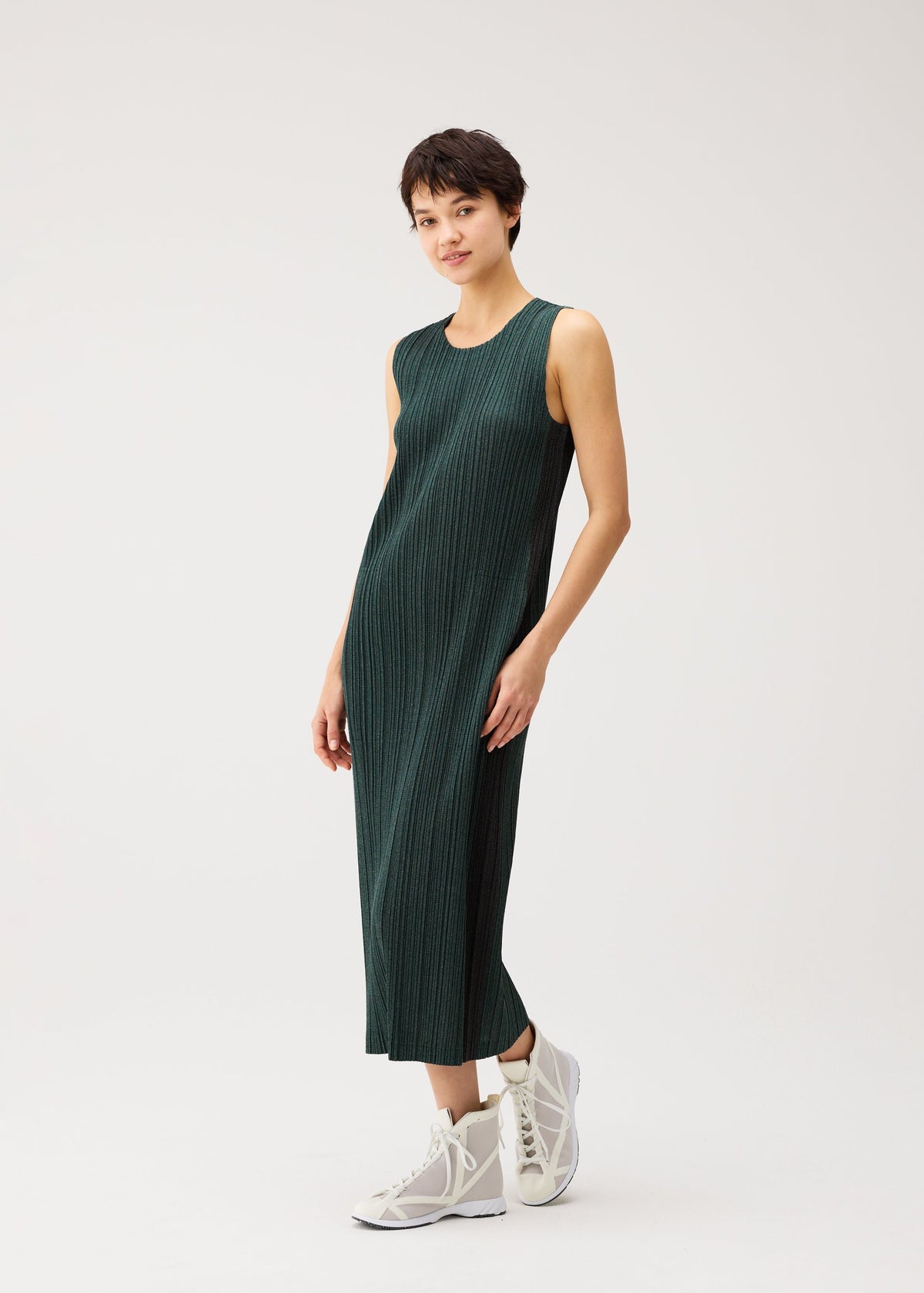 Zoomed view A person wearing the COTTON DENIM by PLEATS PLEASE ISSEY MIYAKE in dark green stands against a white background, styled casually with white high-top sneakers and a slight smile.