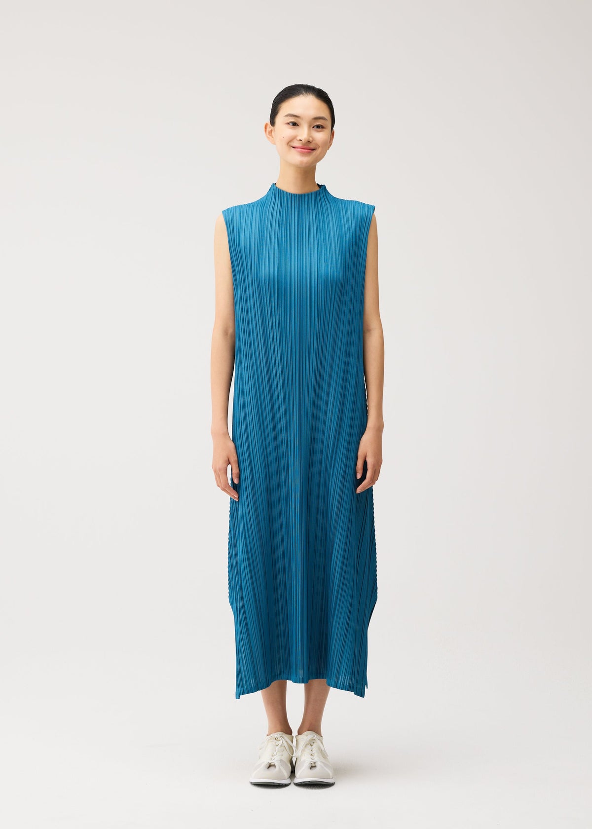 A person stands facing forward, smiling, wearing the PLEATS PLEASE ISSEY MIYAKE MELLOW PLEATS sleeveless bright blue midi dress with pockets and white sneakers, against a plain light background. view 6 of 9
