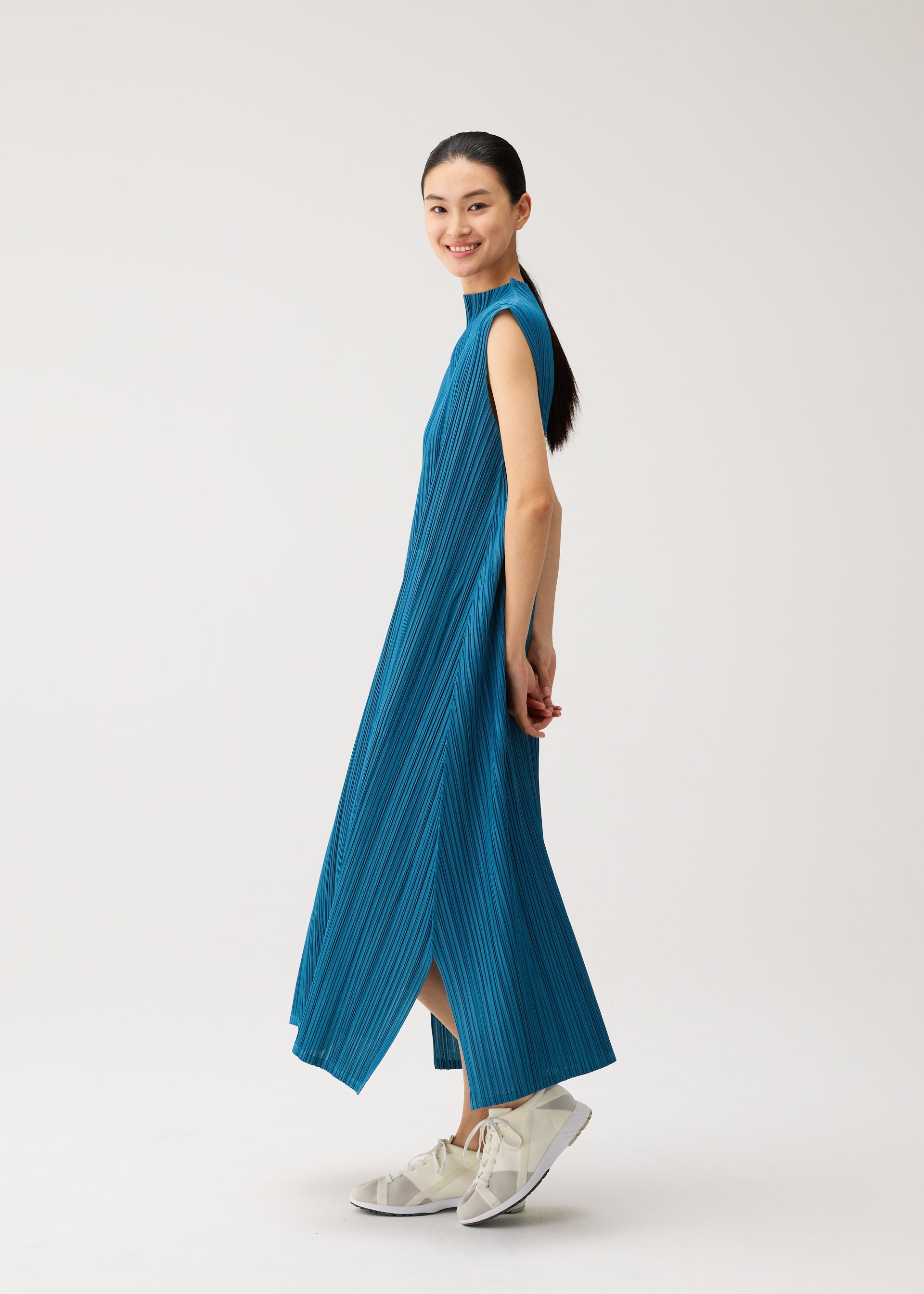 Zoomed view A woman stands sideways and smiles against a plain, light background, wearing the MELLOW PLEATS sleeveless blue textured midi dress with pockets by PLEATS PLEASE ISSEY MIYAKE, paired with white sneakers.
