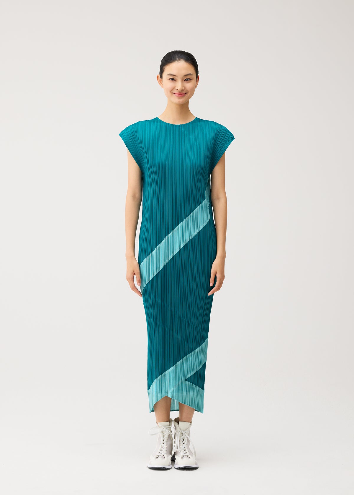 A woman stands smiling in the PLEATS PLEASE ISSEY MIYAKE TWIRL pleated reversible midi dress in teal with light blue diagonal stripes, paired with white high-top sneakers, against a simple background. view 6 of 10