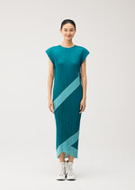 A woman stands smiling in the PLEATS PLEASE ISSEY MIYAKE TWIRL pleated reversible midi dress in teal with light blue diagonal stripes, paired with white high-top sneakers, against a simple background.