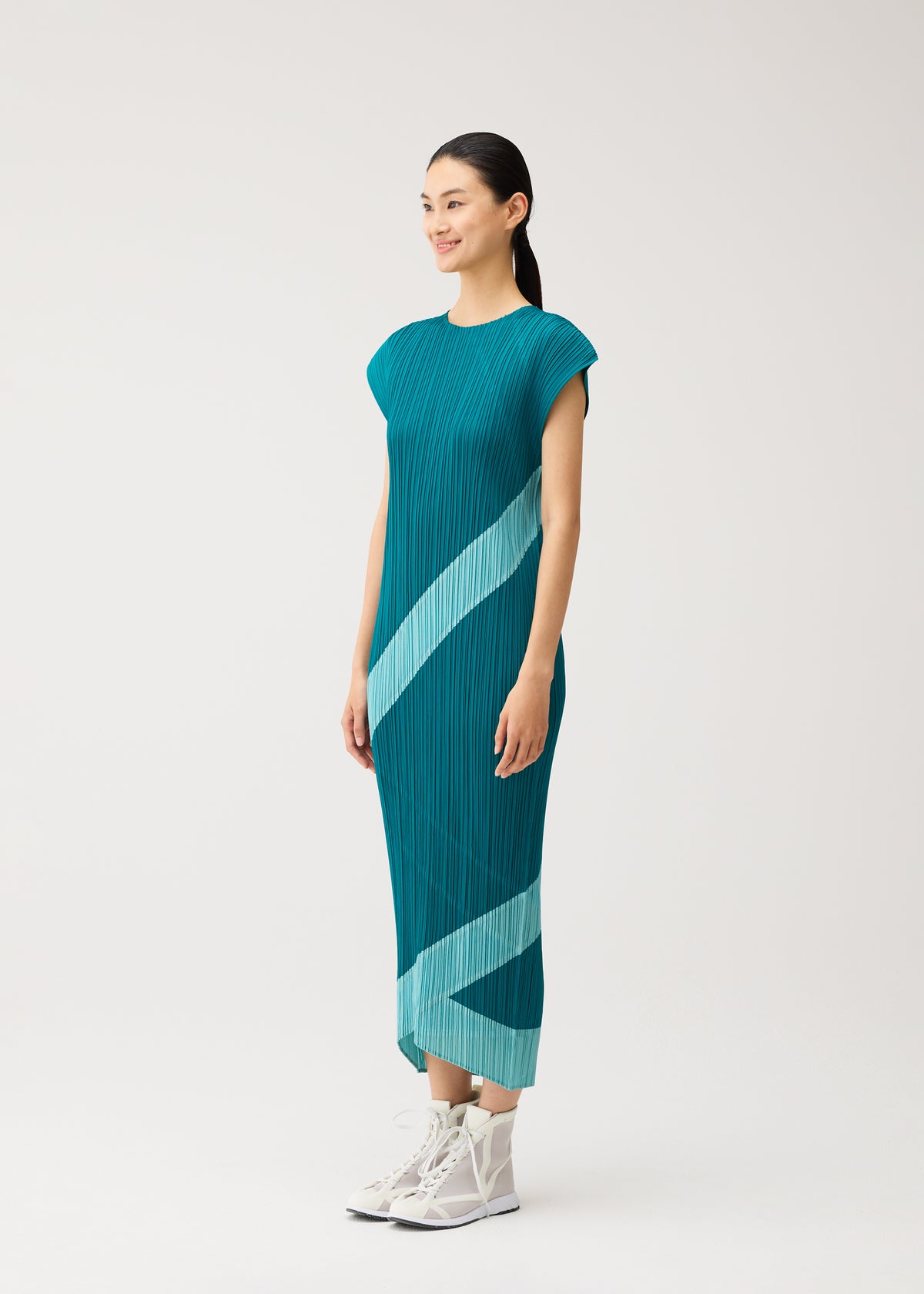 Wearing the TWIRL dress by PLEATS PLEASE ISSEY MIYAKE, a woman stands against a neutral backdrop. The teal midi dress features pleats, sleeveless styling, and light blue diagonal stripes, paired with white high-top sneakers. view 7 of 10