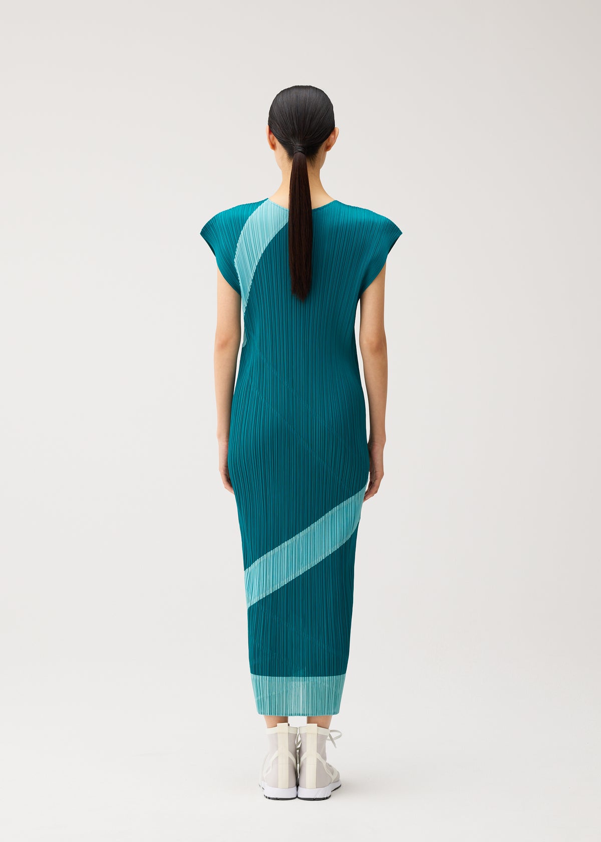 A woman with straight dark hair in a ponytail stands facing away, wearing the TWIRL dress by PLEATS PLEASE ISSEY MIYAKE—a teal, round-neck reversible style featuring light blue diagonal stripes—paired with white shoes, set against a plain light background. view 8 of 10