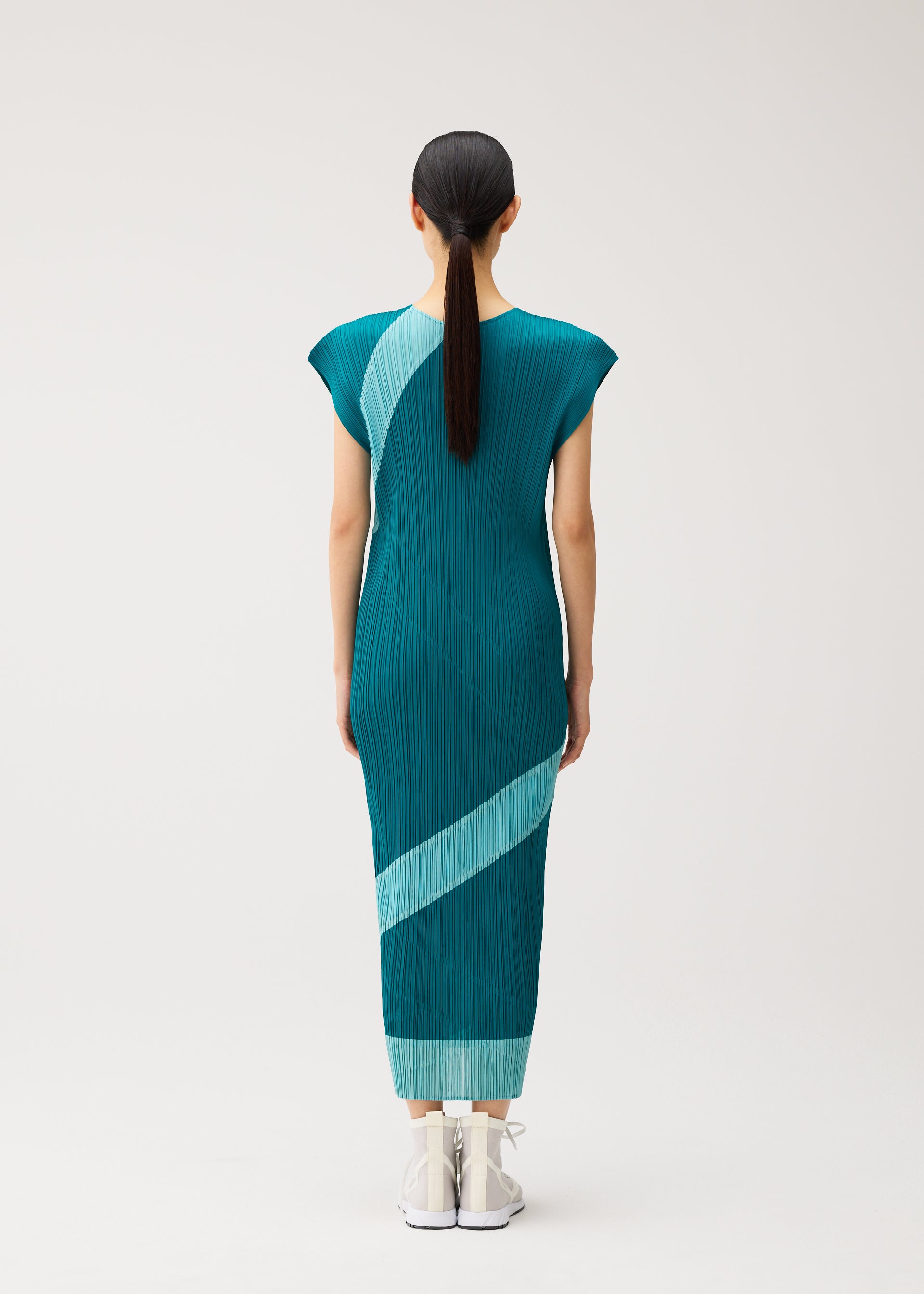 Zoomed view A woman with straight dark hair in a ponytail stands facing away, wearing the TWIRL dress by PLEATS PLEASE ISSEY MIYAKE—a teal, round-neck reversible style featuring light blue diagonal stripes—paired with white shoes, set against a plain light background.