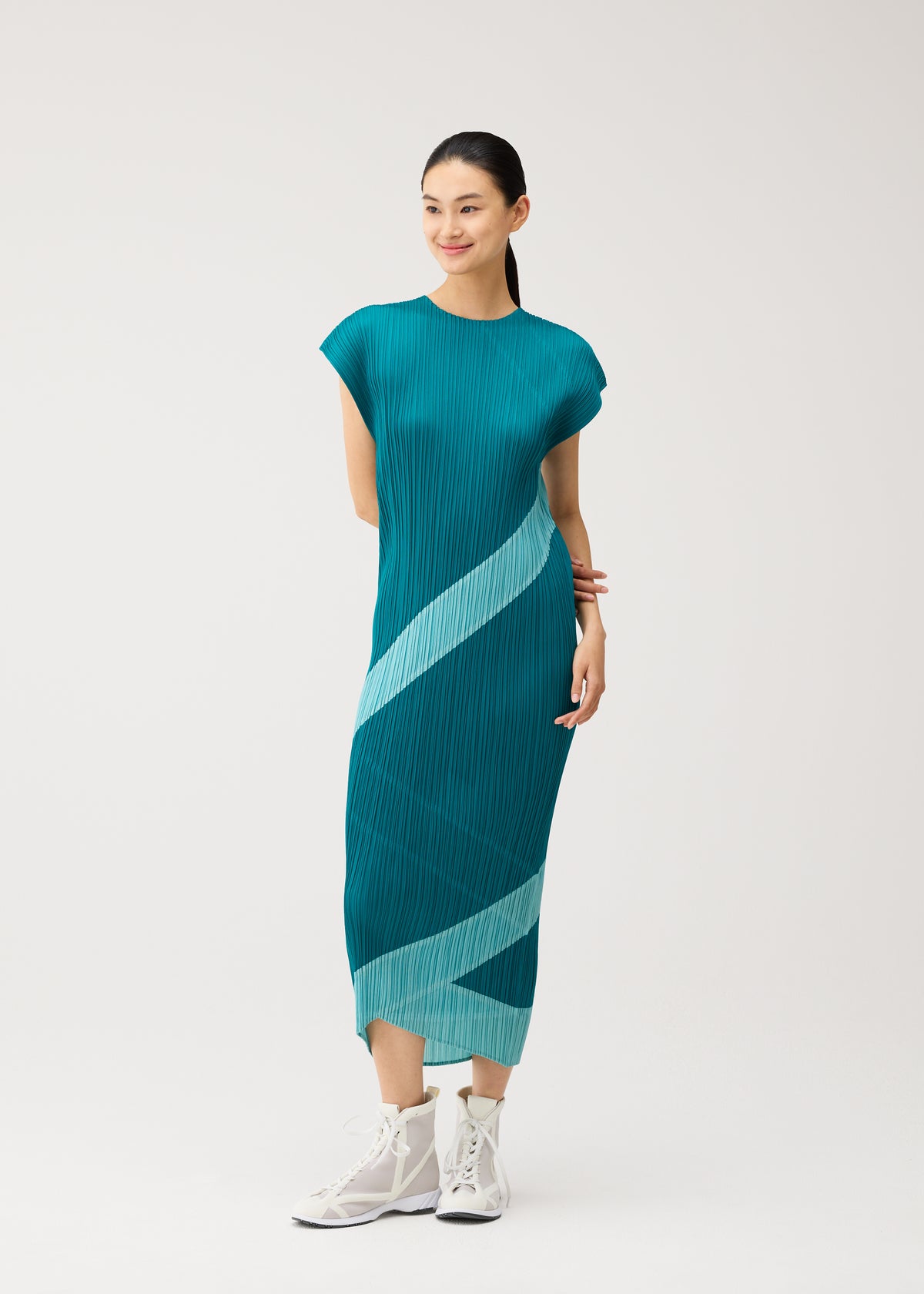 A woman stands smiling with her hands clasped, wearing the PLEATS PLEASE ISSEY MIYAKE TWIRL dress—a teal, pleated round-neck design featuring light blue diagonal stripes—paired with white high-top sneakers against a plain white background. view 9 of 10