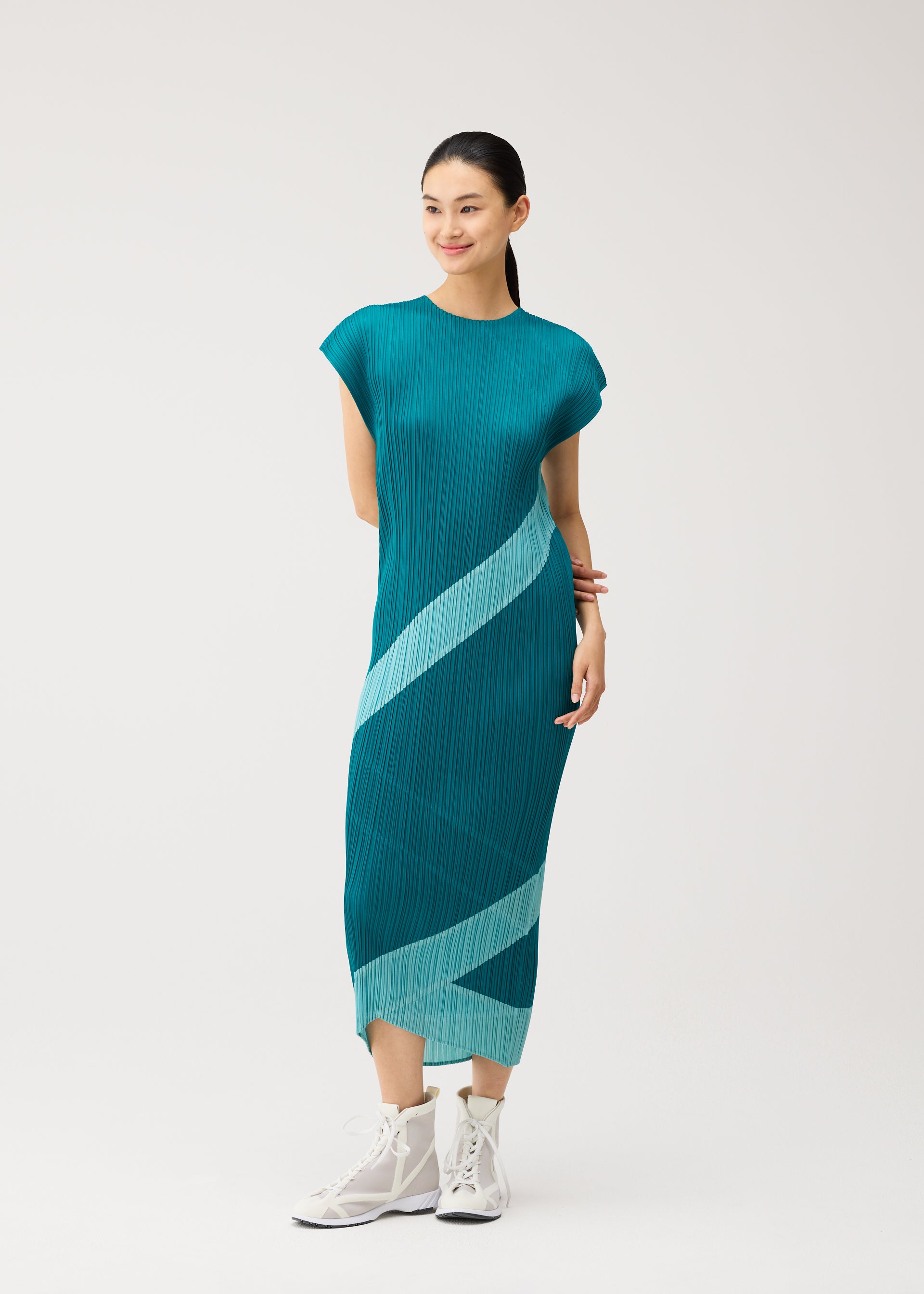 Zoomed view A woman stands smiling with her hands clasped, wearing the PLEATS PLEASE ISSEY MIYAKE TWIRL dress—a teal, pleated round-neck design featuring light blue diagonal stripes—paired with white high-top sneakers against a plain white background.