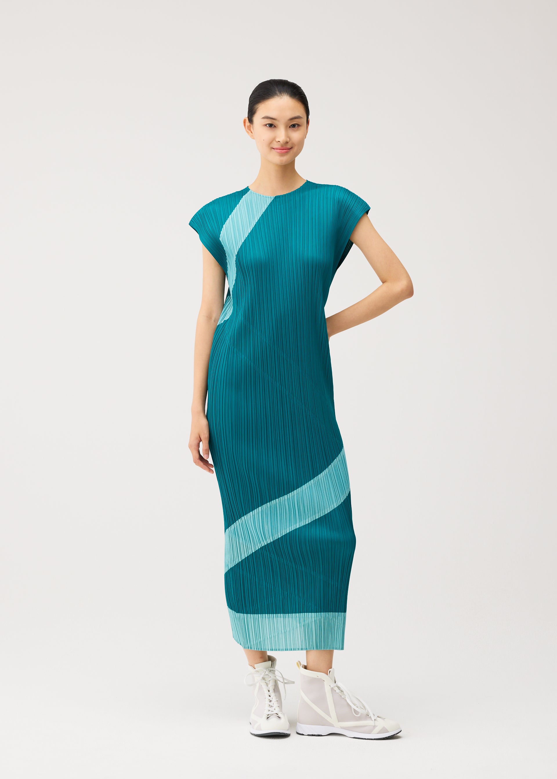 Zoomed view A woman stands against a plain white background, wearing the TWIRL dress by PLEATS PLEASE ISSEY MIYAKE—a teal, round-neck sleeveless dress featuring light blue diagonal stripes—paired with white sneakers and her hair tied back.