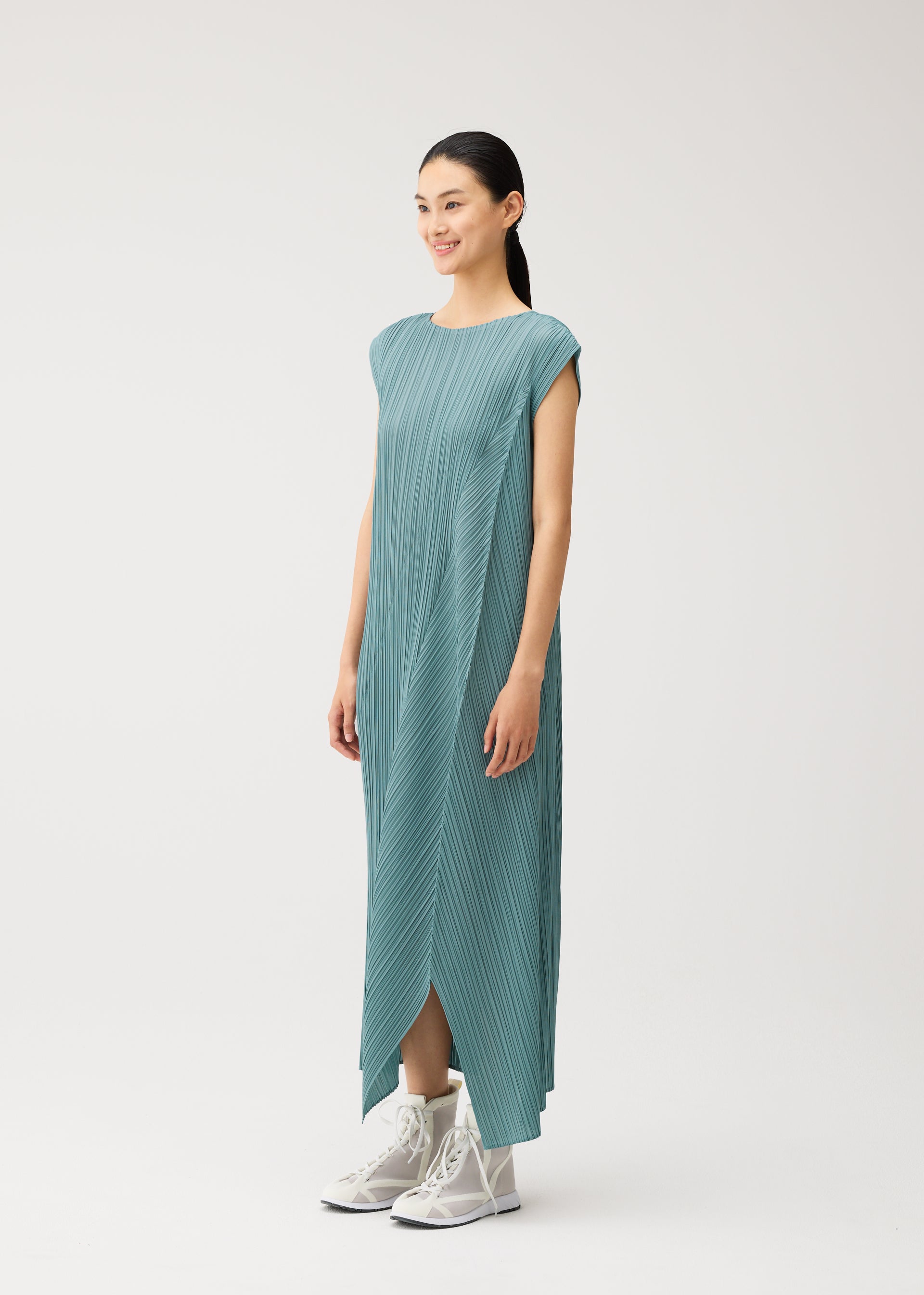 Zoomed view A woman stands smiling in the FOLD teal pleated maxi dress by PLEATS PLEASE ISSEY MIYAKE, featuring a round neckline, cap sleeves, and a layered front. She wears white ankle boots against a plain, light-colored background.