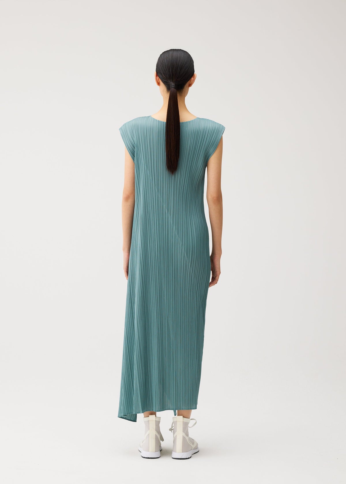 A woman with straight black hair in a low ponytail wears the FOLD sleeveless teal pleated maxi dress with a round neckline from PLEATS PLEASE ISSEY MIYAKE, paired with white sneakers, standing against a plain light background and facing away from the camera. view 7 of 8
