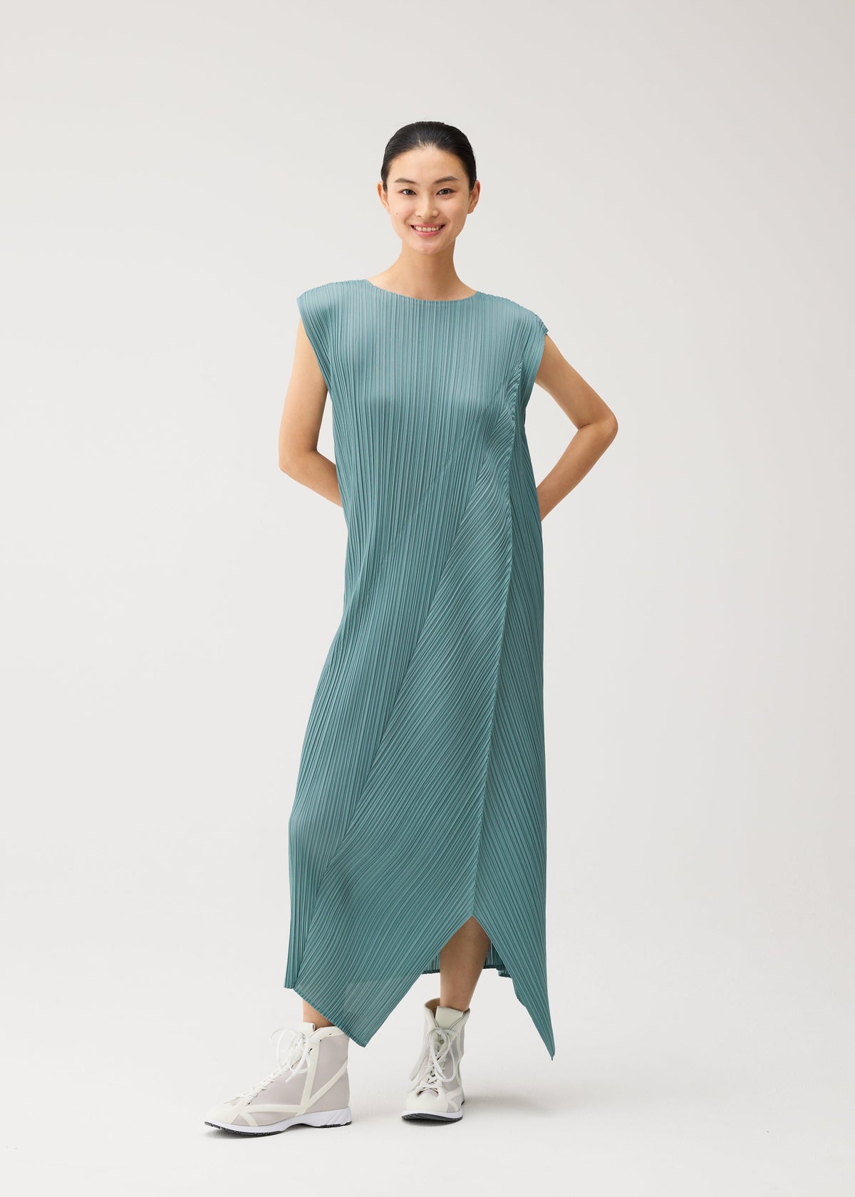 A person stands smiling against a plain light background, wearing the FOLD dress by PLEATS PLEASE ISSEY MIYAKE—a teal, pleated, sleeveless piece with a round neck and an asymmetrical hem—paired with white high-top sneakers for a modern, casually elegant look. view 8 of 8