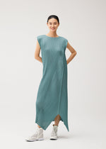 A person stands smiling against a plain light background, wearing the FOLD dress by PLEATS PLEASE ISSEY MIYAKE—a teal, pleated, sleeveless piece with a round neck and an asymmetrical hem—paired with white high-top sneakers for a modern, casually elegant look.
