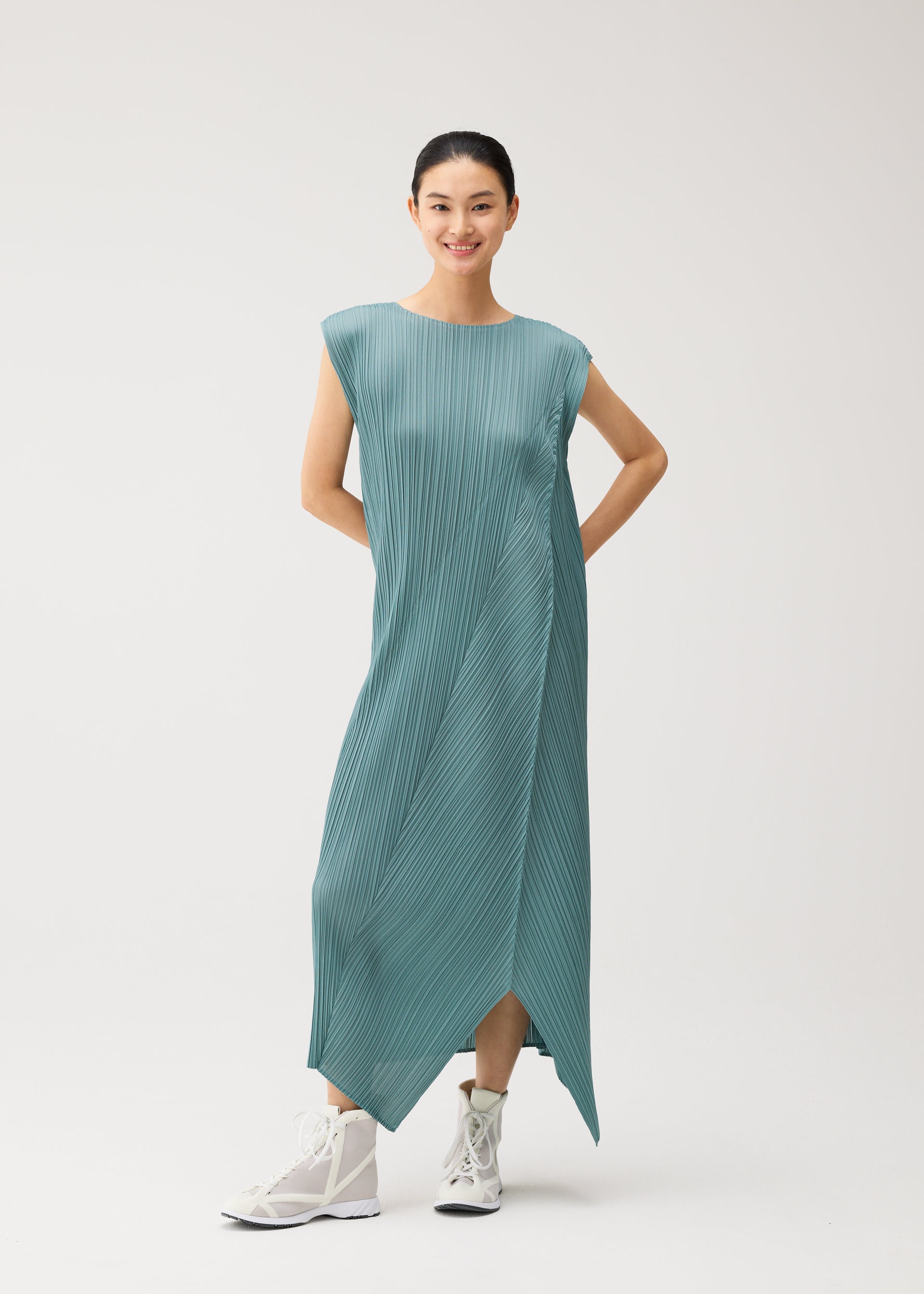 Zoomed view A person stands smiling against a plain light background, wearing the FOLD dress by PLEATS PLEASE ISSEY MIYAKE—a teal, pleated, sleeveless piece with a round neck and an asymmetrical hem—paired with white high-top sneakers for a modern, casually elegant look.
