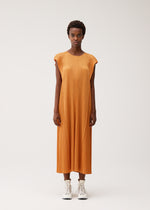 A person stands against a plain background wearing the BOUQUET dress by PLEATS PLEASE ISSEY MIYAKE—an orange, loose-fitting, sleeveless round-neck pleated dress falling below the knees—paired with white high-top sneakers.