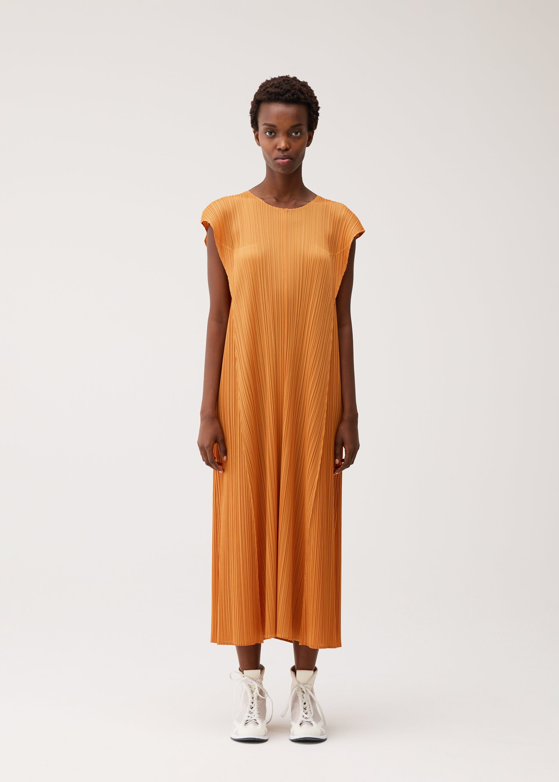 Zoomed view A person stands against a plain background wearing the BOUQUET dress by PLEATS PLEASE ISSEY MIYAKE—an orange, loose-fitting, sleeveless round-neck pleated dress falling below the knees—paired with white high-top sneakers.