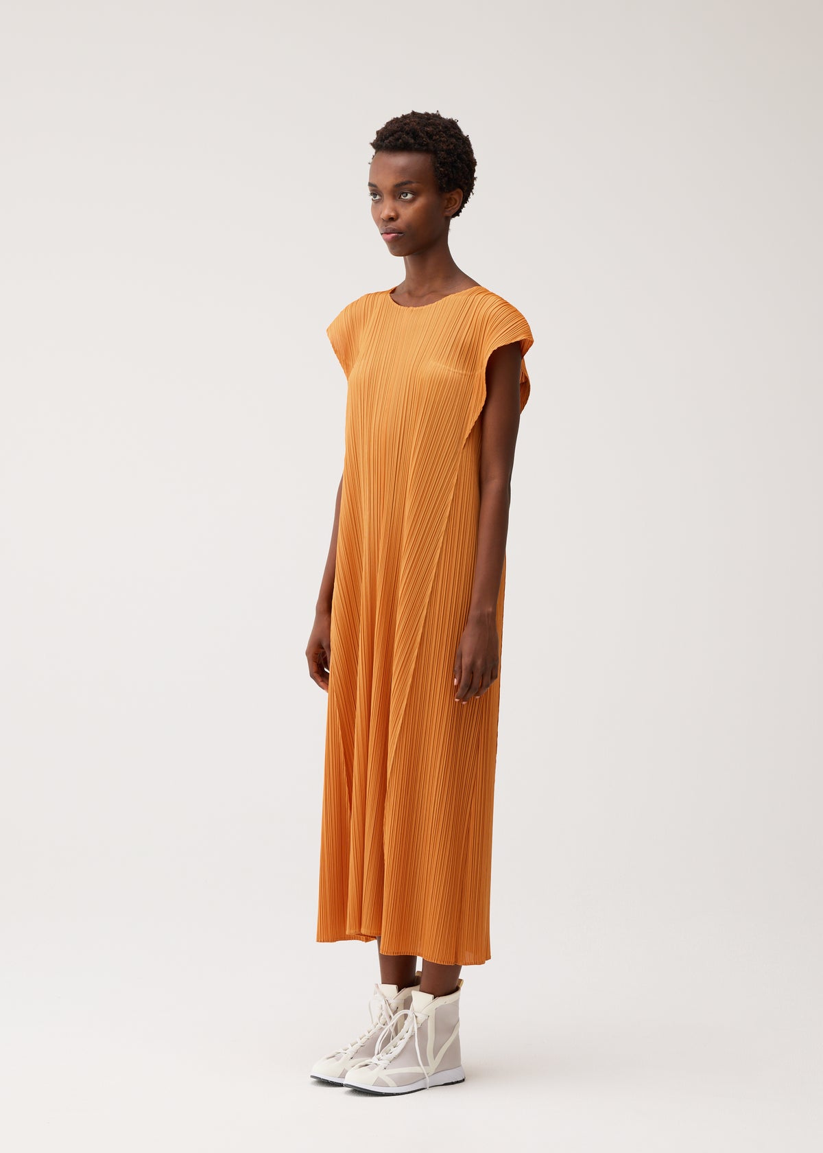A person wears the BOUQUET dress by PLEATS PLEASE ISSEY MIYAKE, a floral-inspired loose-fitting orange piece with cap sleeves, paired with white high-top sneakers against a plain background. view 7 of 10