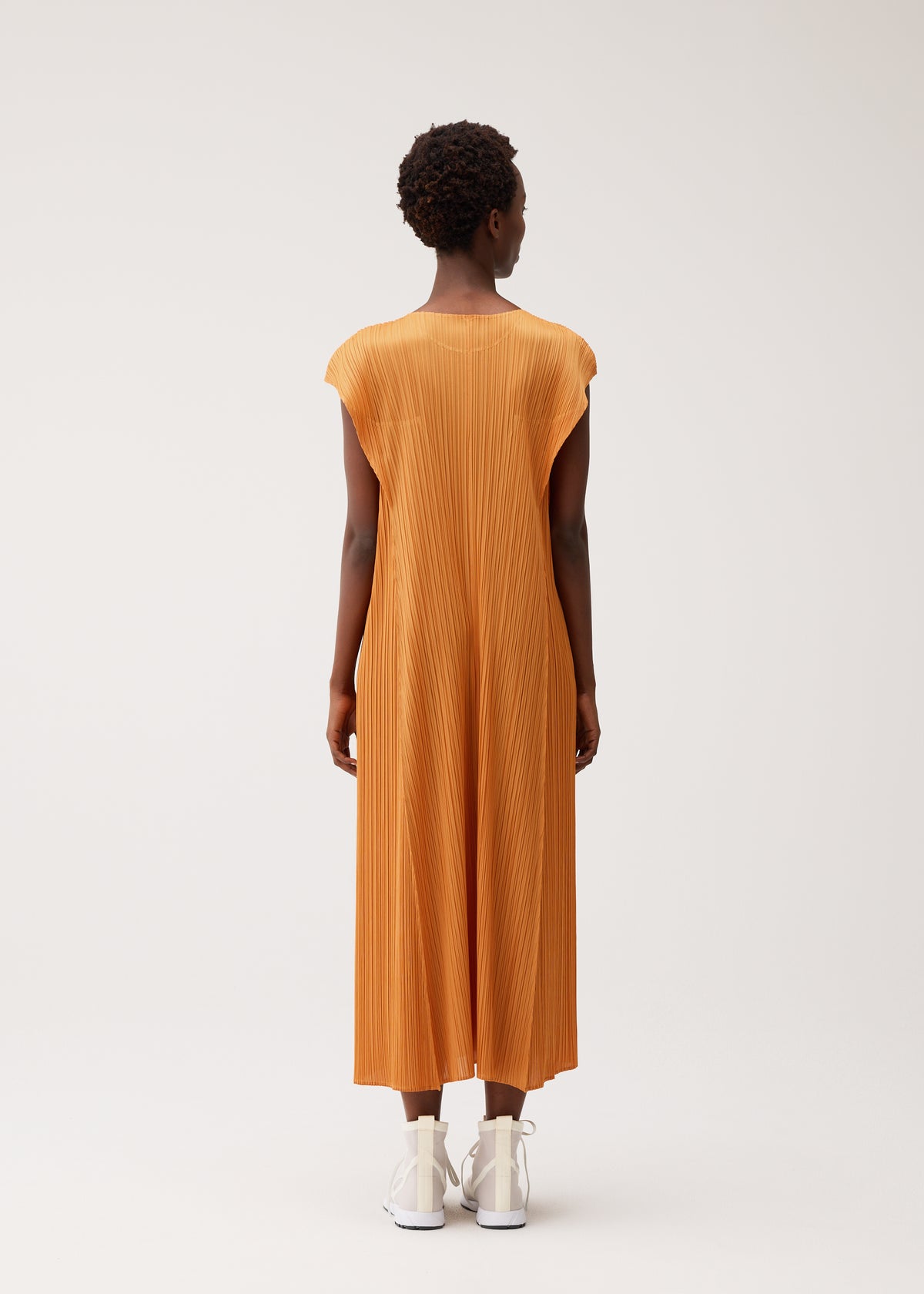 A person wearing the BOUQUET orange midi dress by PLEATS PLEASE ISSEY MIYAKE with white shoes faces away from the camera against a plain, light background. view 8 of 10