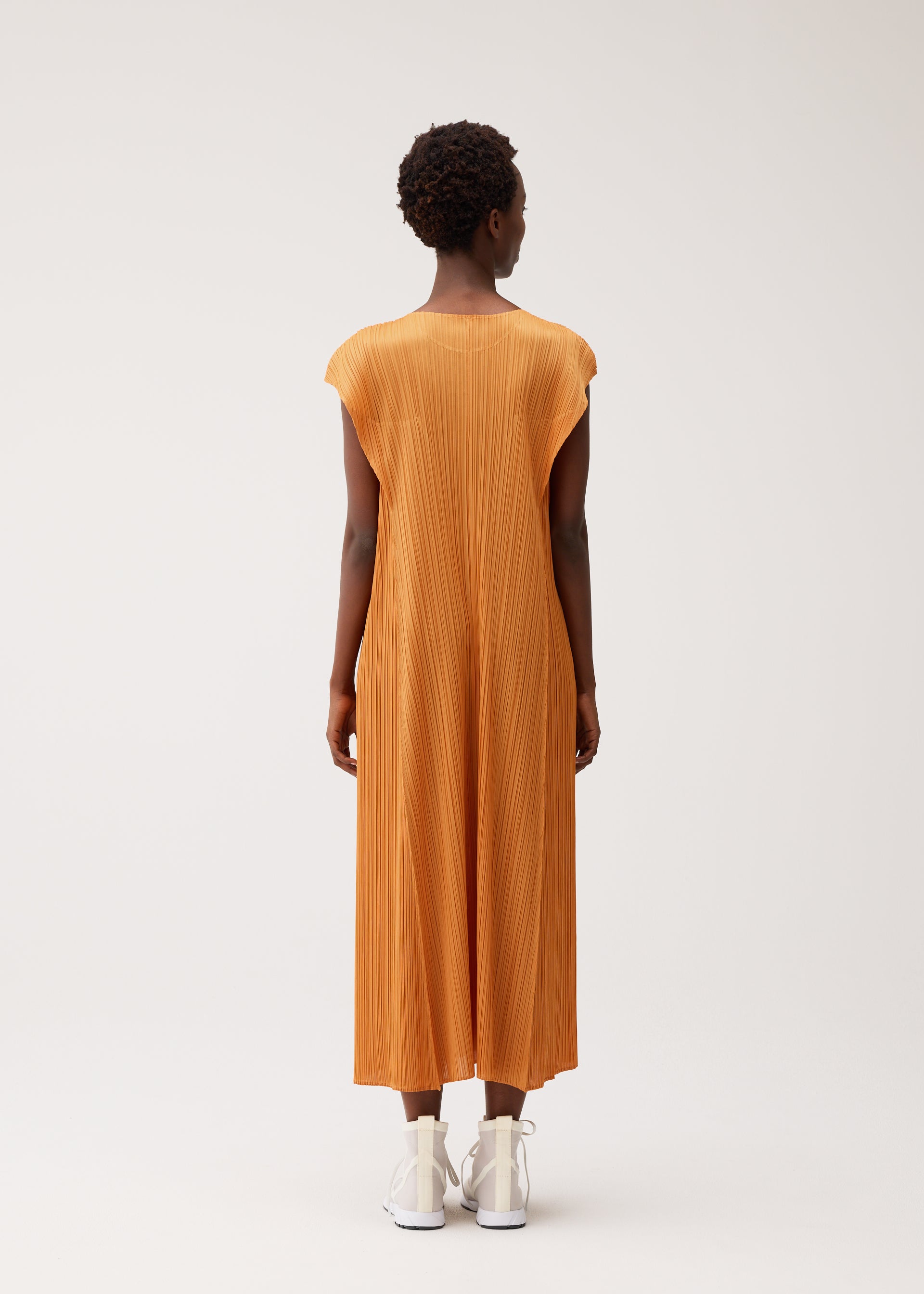 Zoomed view A person wearing the BOUQUET orange midi dress by PLEATS PLEASE ISSEY MIYAKE with white shoes faces away from the camera against a plain, light background.
