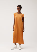 A person in the BOUQUET pleated midi dress by PLEATS PLEASE ISSEY MIYAKE, featuring a loose orange floral design with cap sleeves, paired with white high-top sneakers, stands smiling against a plain light background.