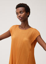A woman in the BOUQUET pleated orange dress by PLEATS PLEASE ISSEY MIYAKE stands with arms slightly outstretched, gazing thoughtfully to the side against a plain light background.