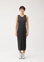A person in the COTTON DENIM BLACK dress by PLEATS PLEASE ISSEY MIYAKE stands smiling against a plain white background, styled with white sneakers.