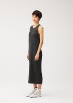 A woman poses sideways in the COTTON DENIM BLACK dress by PLEATS PLEASE ISSEY MIYAKE—an ankle-length, sleeveless gray piece with a denim look—paired with white high-top sneakers against a white background.