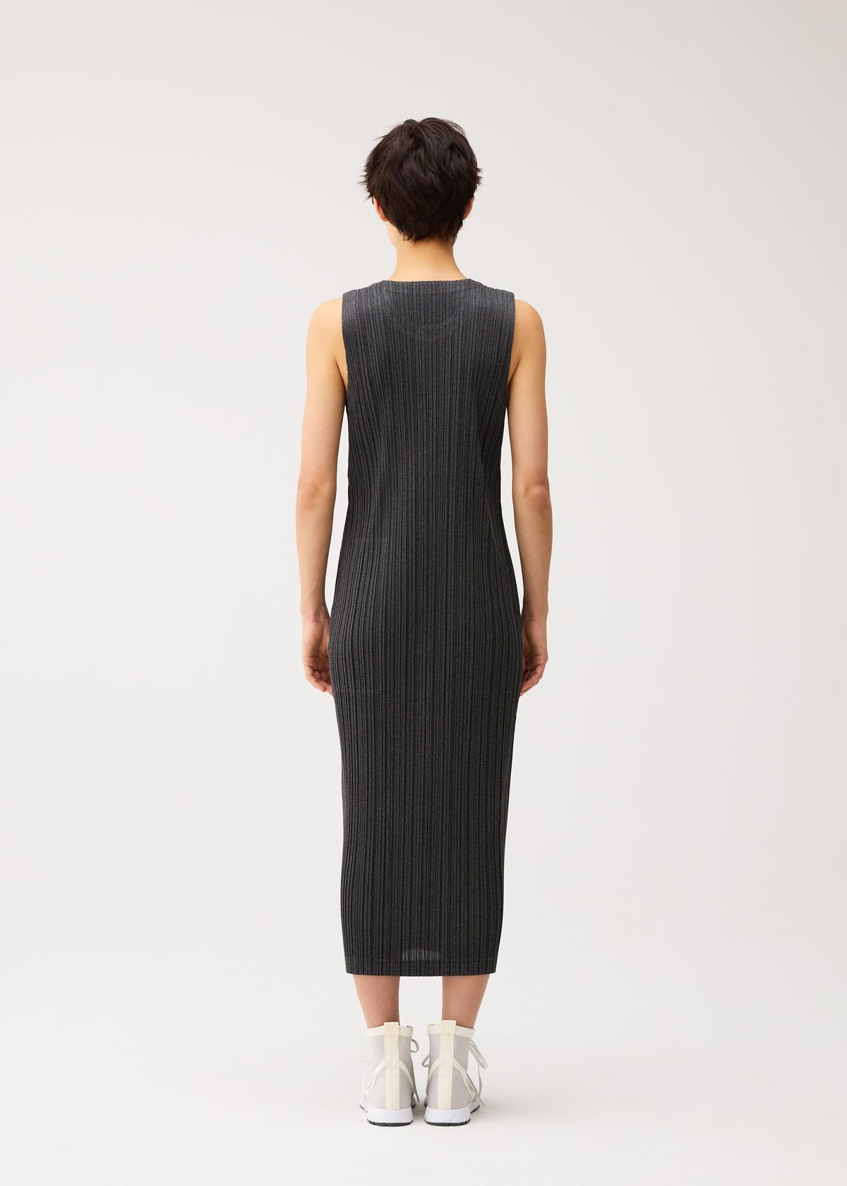 Zoomed view A person with short hair faces away, wearing the PLEATS PLEASE ISSEY MIYAKE COTTON DENIM BLACK sleeveless midi dress and white high-top sneakers against a plain light background.