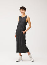 A person with short dark hair wears the PLEATS PLEASE ISSEY MIYAKE COTTON DENIM BLACK, a sleeveless, ankle-length dress, with white high-top sneakers, standing and smiling with one hand in their pocket.