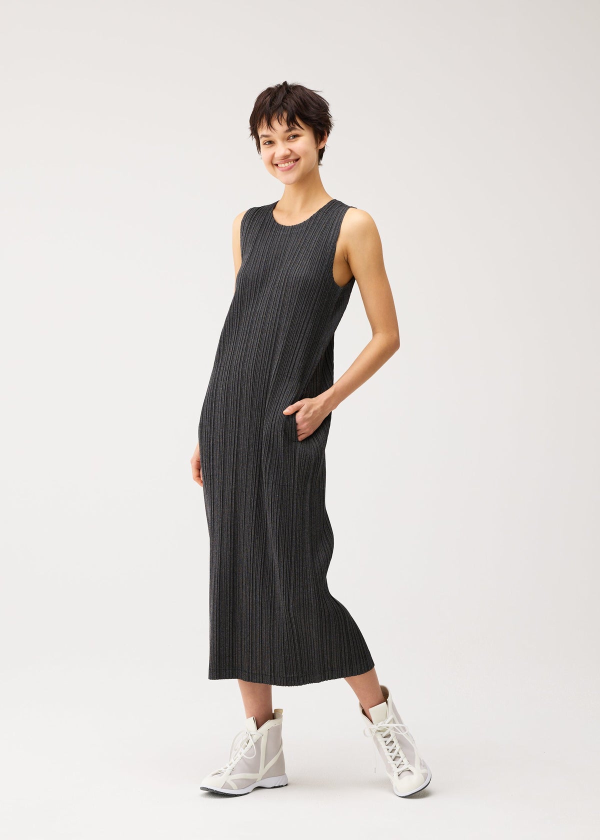 Zoomed view A person with short dark hair wears the PLEATS PLEASE ISSEY MIYAKE COTTON DENIM BLACK, a sleeveless, ankle-length dress, with white high-top sneakers, standing and smiling with one hand in their pocket.