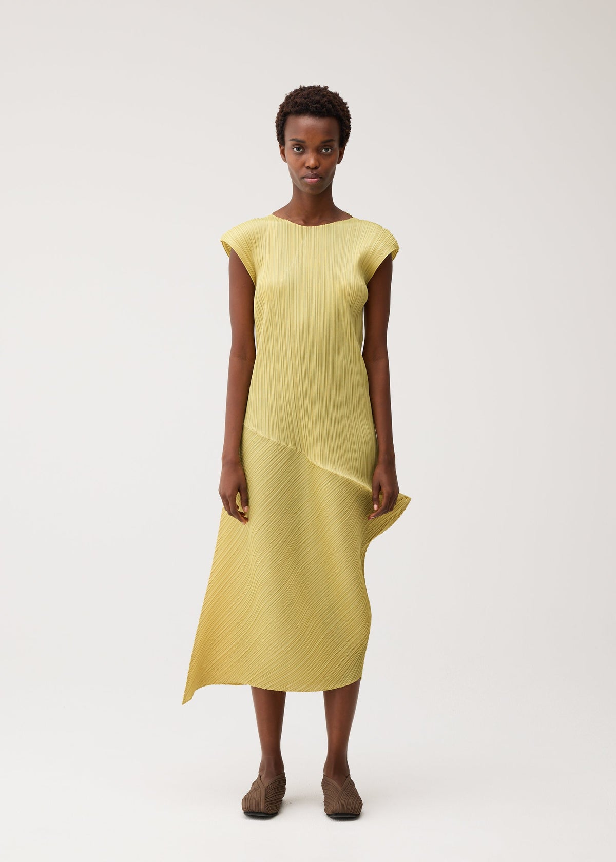 A woman stands against a plain background, wearing the LEAP dress by PLEATS PLEASE ISSEY MIYAKE—yellow, pleated, round-neck, with an asymmetrical design and brown shoes—looking directly at the camera. view 5 of 8