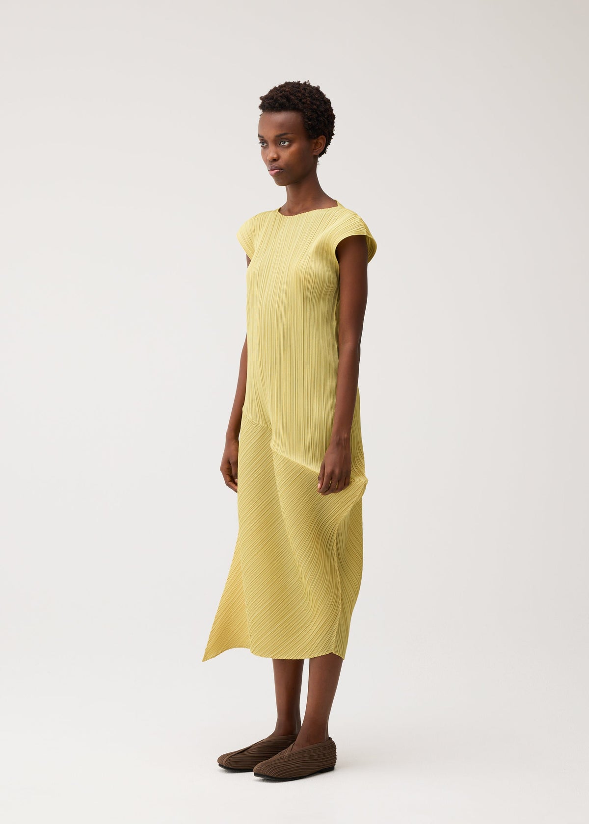 A woman in a studio wears the LEAP pleated midi dress in light yellow by PLEATS PLEASE ISSEY MIYAKE, paired with brown shoes, gazing slightly left with a neutral expression. view 6 of 8