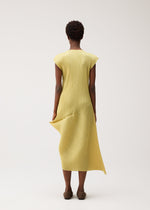 A person stands facing away, wearing the LEAP dress by PLEATS PLEASE ISSEY MIYAKE—a pleated, light yellow, round-neck dress with short sleeves and brown shoes against a plain background.