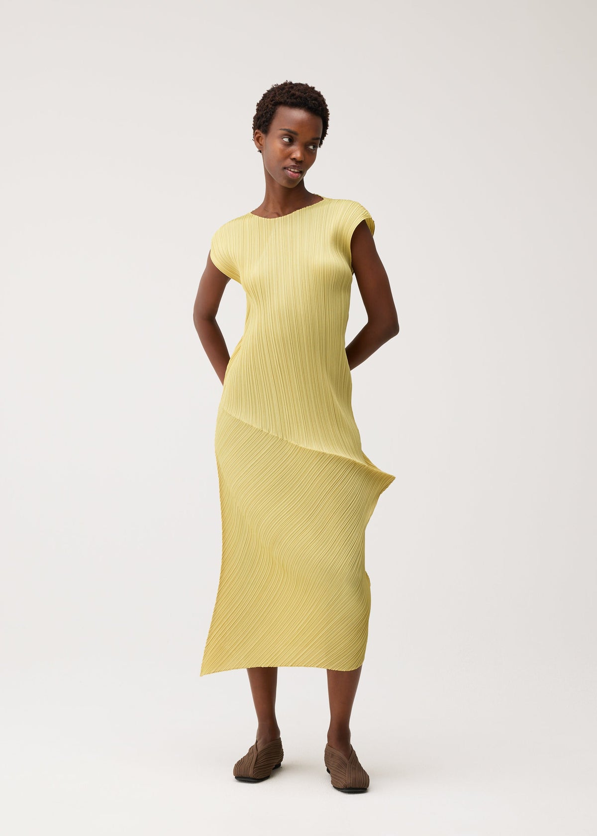 Zoomed view A woman poses against a plain light background, hands behind her back, wearing the LEAP short-sleeved yellow dress by PLEATS PLEASE ISSEY MIYAKE and brown flat shoes.