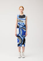 A woman with light skin and red hair stands facing forward, wearing the ROLL dress by PLEATS PLEASE ISSEY MIYAKE—a sleeveless, knee-length piece in blue, black, and white abstract print—with white ankle boots against a plain light background.