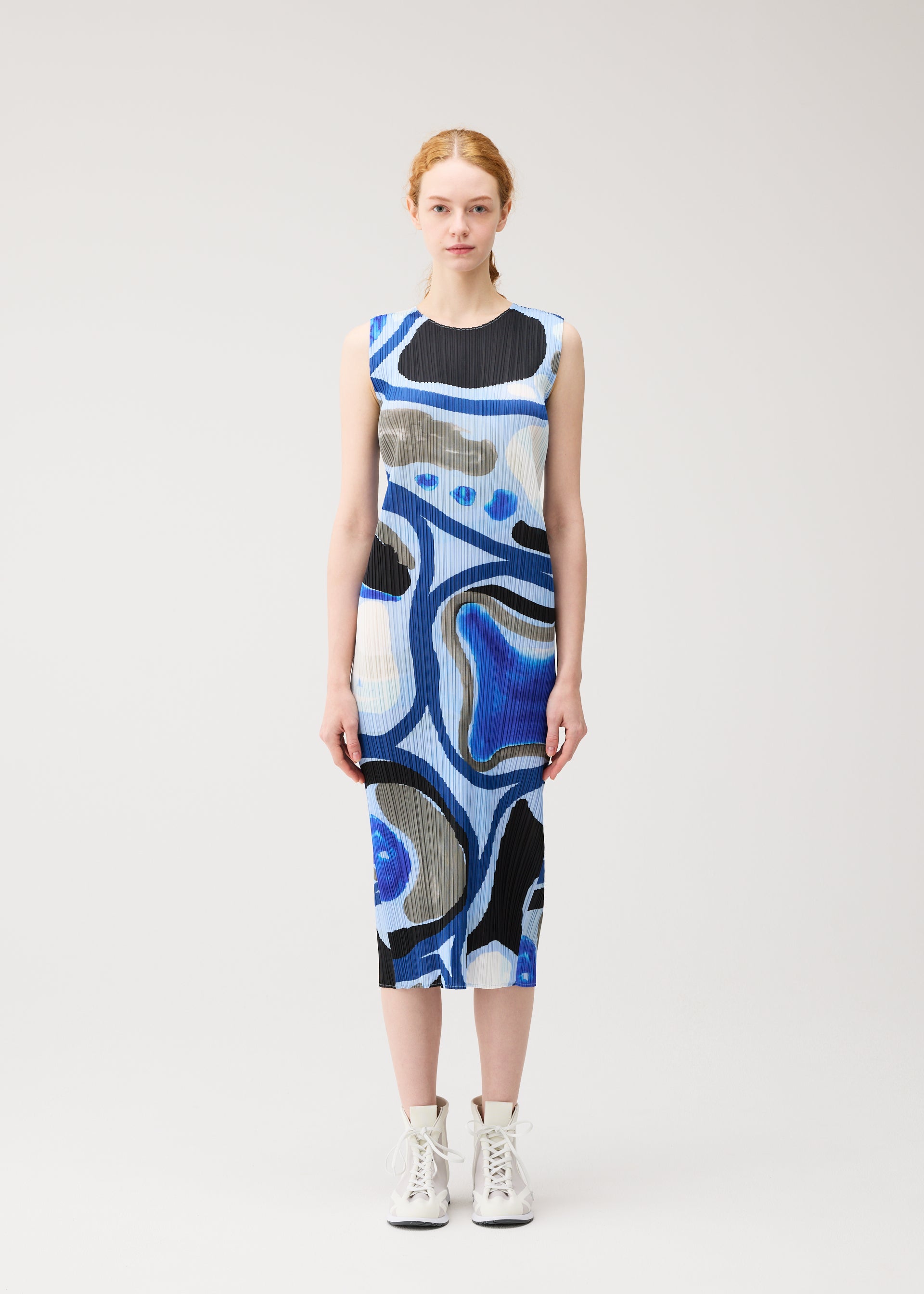 Zoomed view A woman with light skin and red hair stands facing forward, wearing the ROLL dress by PLEATS PLEASE ISSEY MIYAKE—a sleeveless, knee-length piece in blue, black, and white abstract print—with white ankle boots against a plain light background.