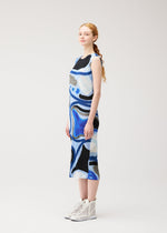 A woman with long red hair in a ponytail stands sideways against a light background, wearing the ROLL sleeveless knee-length dress with blue, black, and white abstract prints by PLEATS PLEASE ISSEY MIYAKE and white high-top shoes.