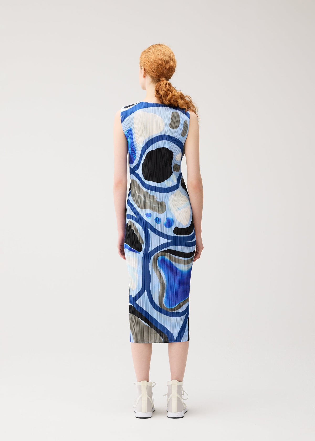 A person with long, curly red hair in a ponytail stands facing away, wearing the PLEATS PLEASE ISSEY MIYAKE ROLL sleeveless, knee-length printed dress with an abstract blue, black, and white pattern and white high-top shoes against a plain background. view 6 of 7