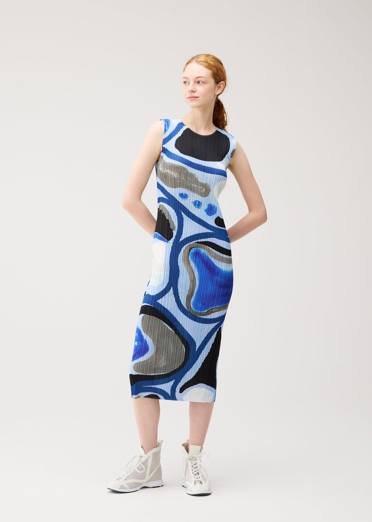 A woman with red hair wears PLEATS PLEASE ISSEY MIYAKE's ROLL, a sleeveless, mid-length dress with bold blue, black, beige, and white abstract prints. She stands with hands in pockets and looks to the side in white high-top sneakers. view 7 of 7