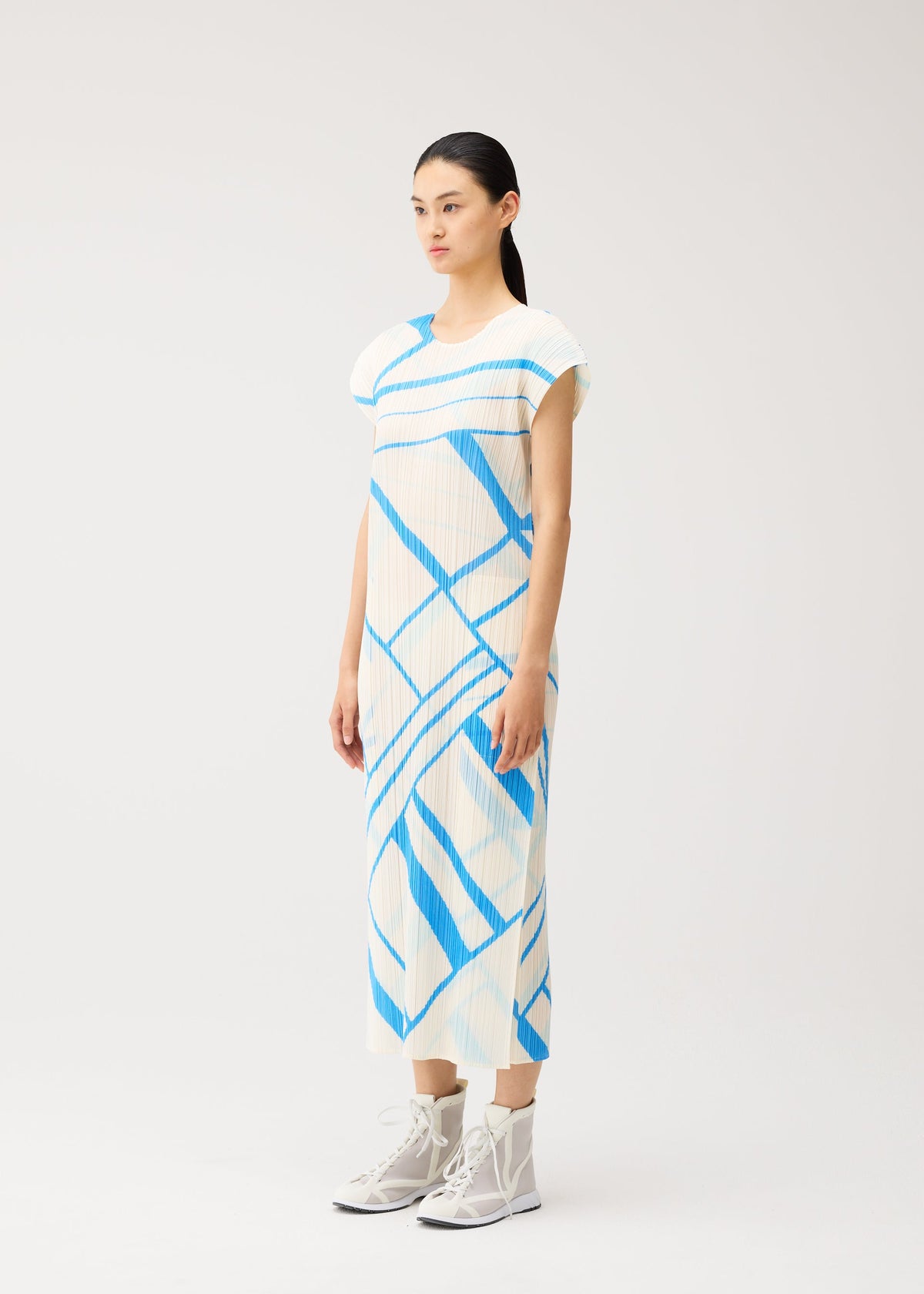 A woman stands against a plain light background, dressed in the FOLD LINE dress by PLEATS PLEASE ISSEY MIYAKE. The white round-neck dress features blue diagonal stripes and French sleeves, paired with white high-top shoes. Its minimalist design exudes effortless elegance. view 6 of 8