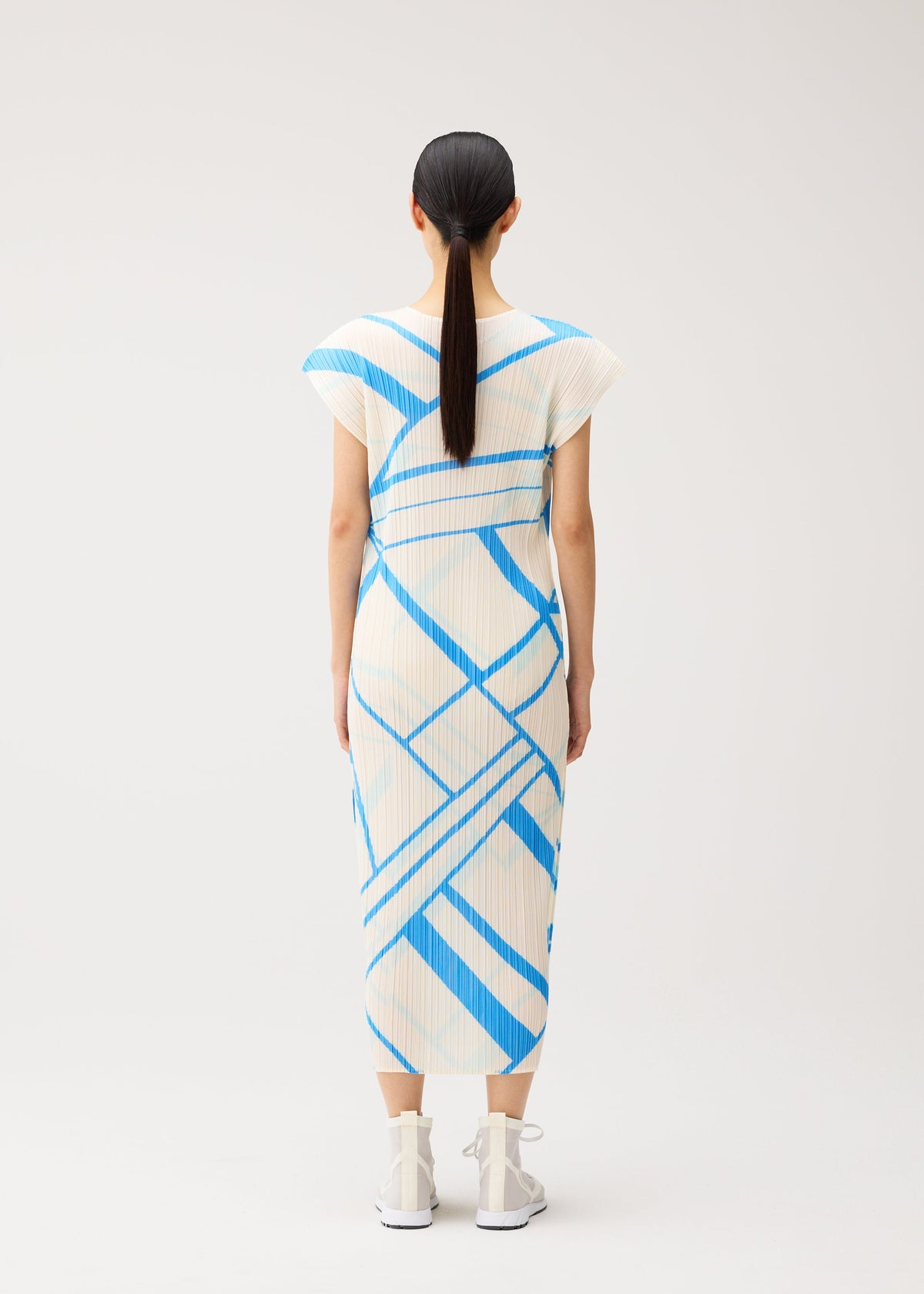 A woman stands with her back to the viewer, wearing the FOLD LINE midi dress by PLEATS PLEASE ISSEY MIYAKE—a white dress featuring blue geometric stripes and French sleeves. She pairs it with white socks and white sneakers, and her straight dark hair is tied in a low ponytail. The plain, light-colored background highlights the minimalist design of her ensemble. view 7 of 8